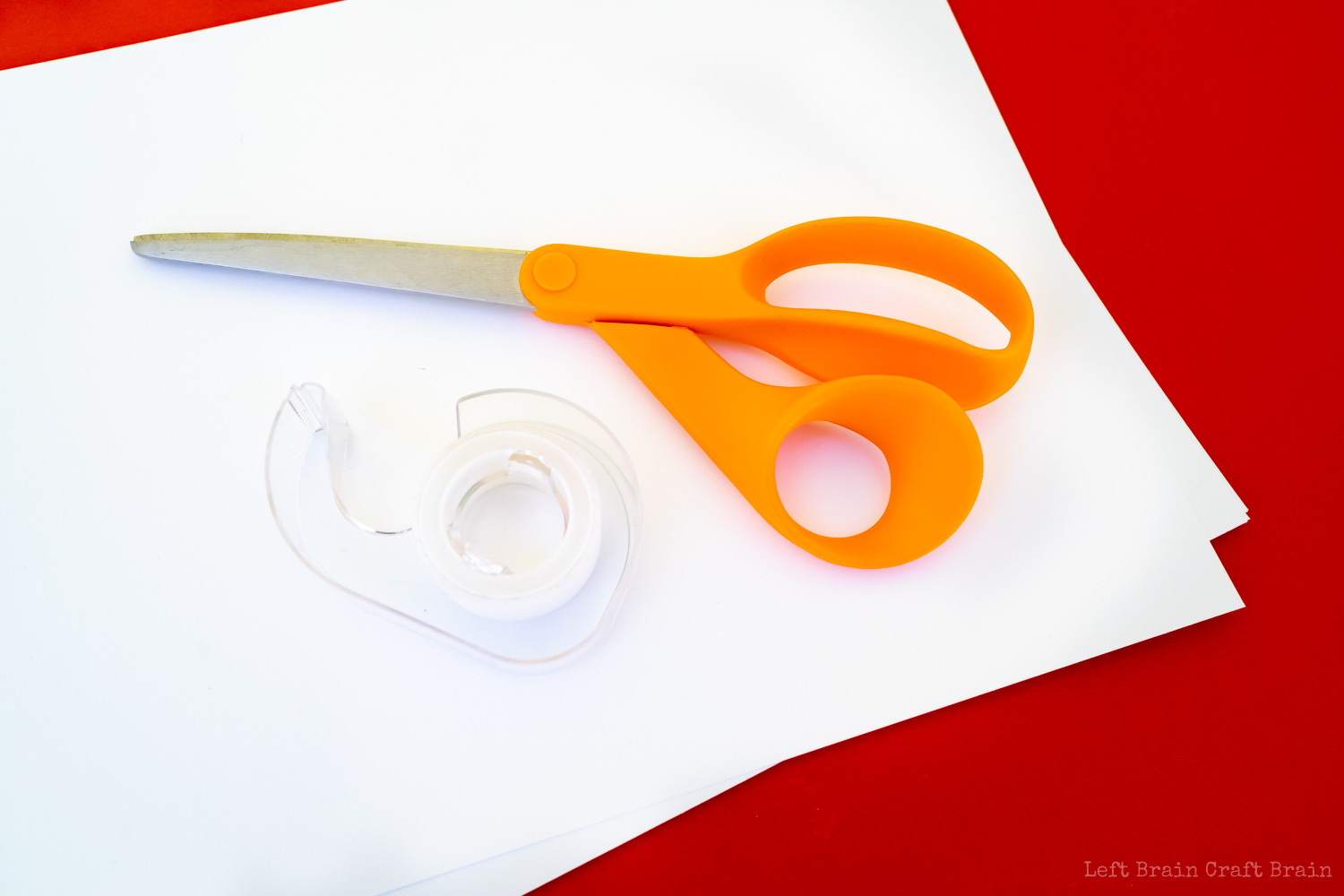 white paper, orange scissors, and tape on a red background