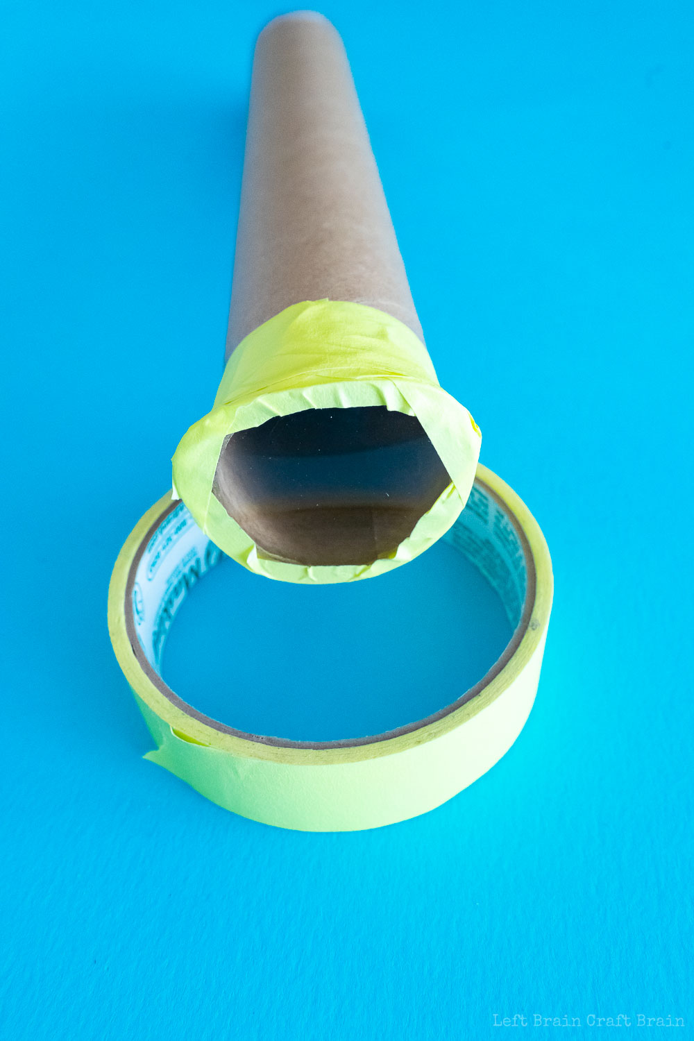 how to make a telescope - End view of paper towel roll with lens taped to end with yellow masking tape. Roll is sitting on yellow tape roll on blue paper