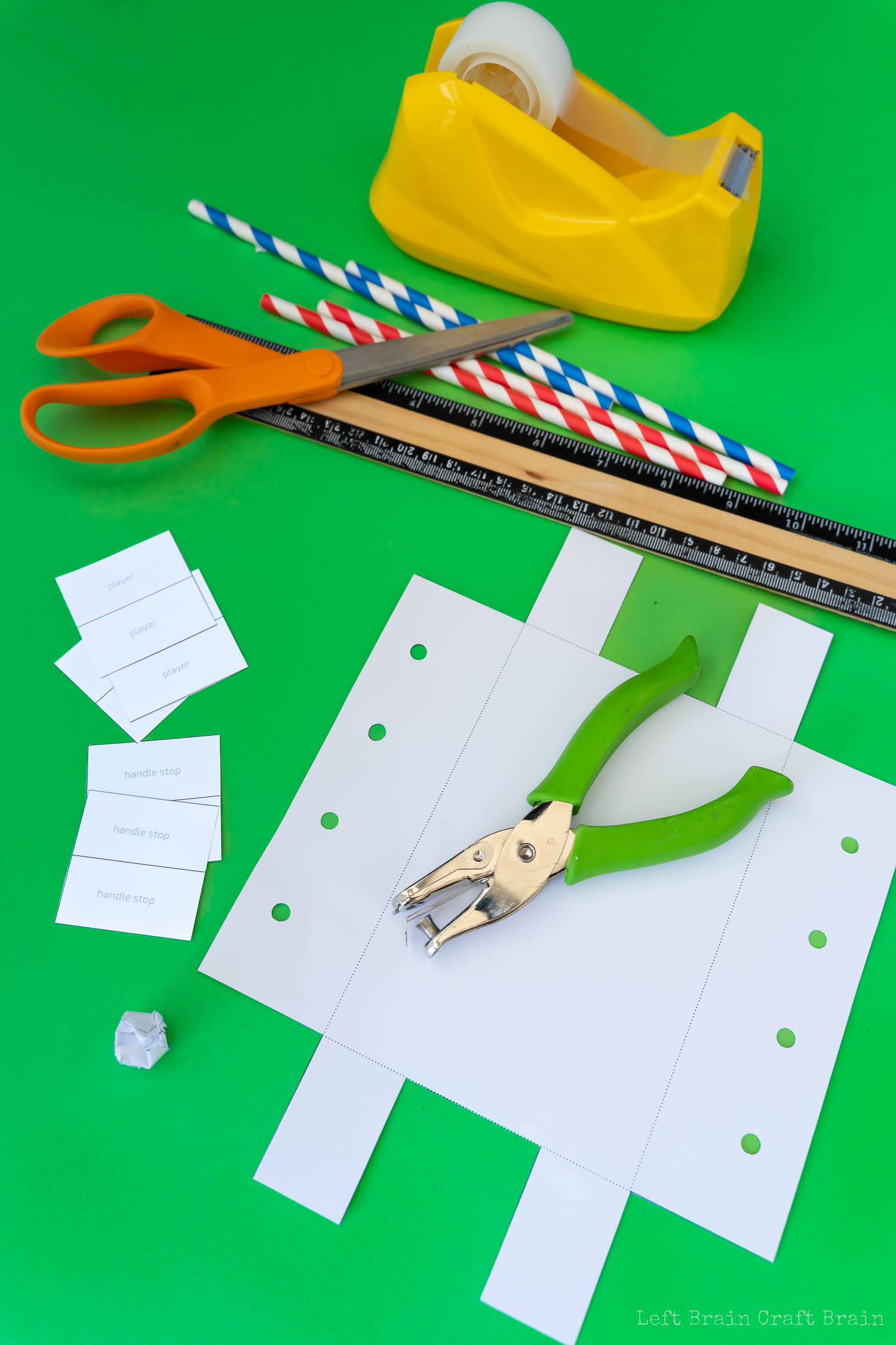 cut and punch holes in paper foosball template (green hole punch, red and blue striped paper straws, orange-handled scissors, yellow tape dispenser, black and wood ruler, white paper template, crumpled paper ball, small pieces of paper that say handle stop and player on green paper background)