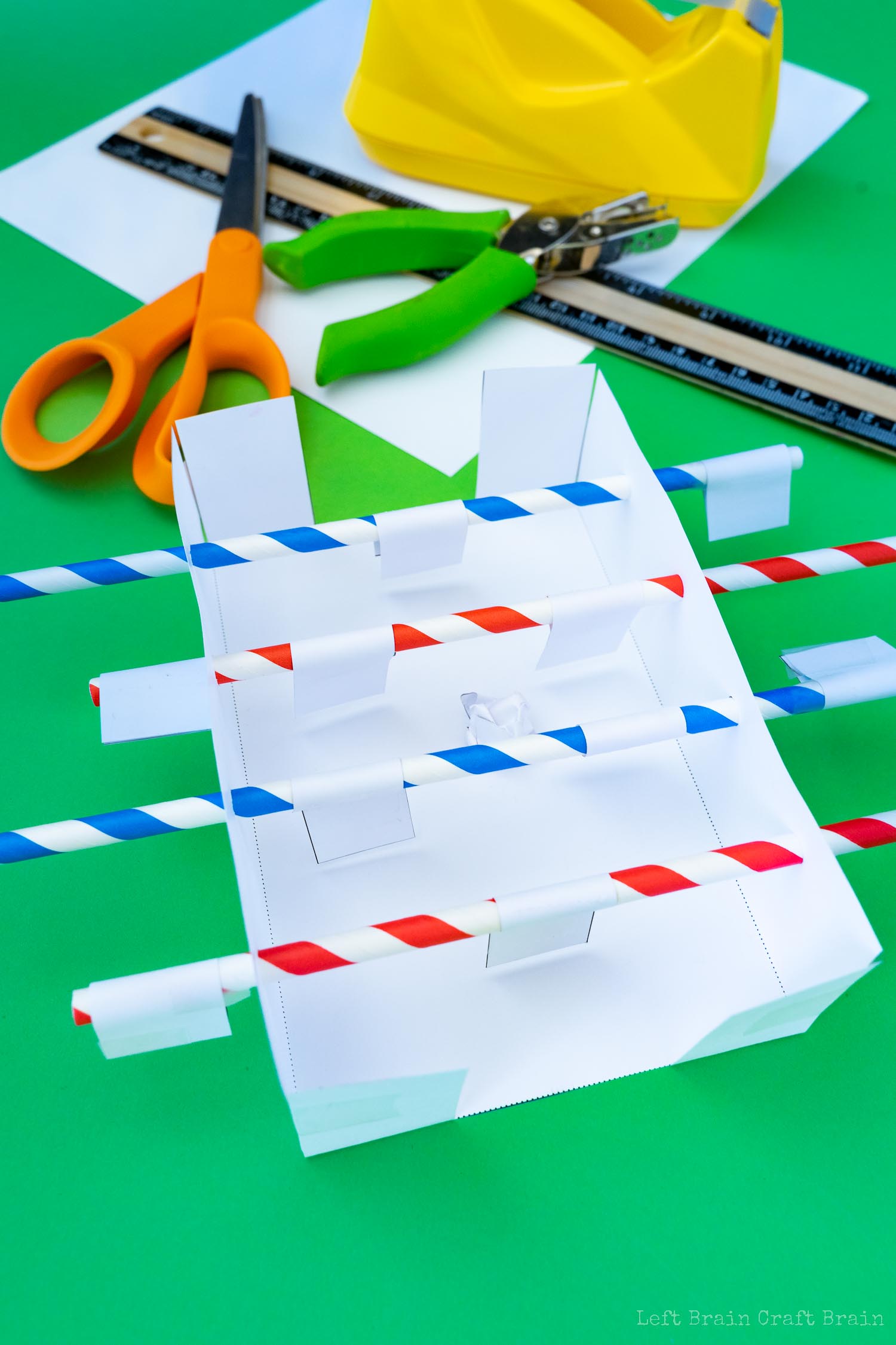 Finished paper foosball table with ball on field (green hole punch, red and blue striped paper straws, orange-handled scissors, yellow tape dispenser, black and wood ruler, white paper template, crumpled paper ball, small pieces of paper that say player on green paper background)