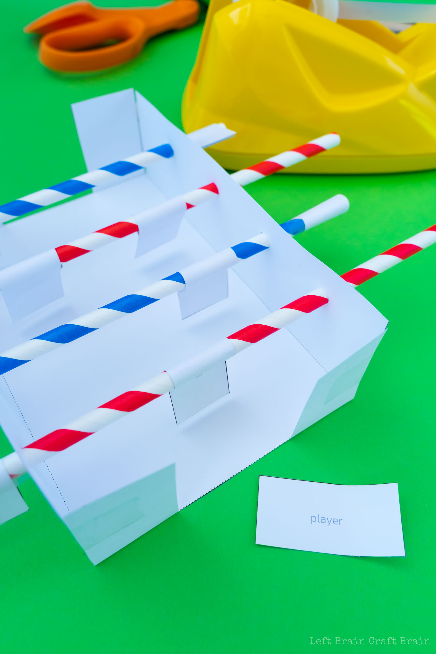 Fold player papers around center of straws (green hole punch, red and blue striped paper straws, orange-handled scissors, yellow tape dispenser, black and wood ruler, white paper template, crumpled paper ball, small pieces of paper that say player on green paper background)