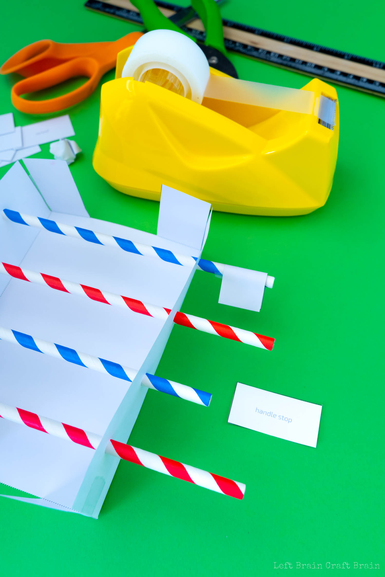 Fold handle stop paper on end of straw (green hole punch, red and blue striped paper straws, orange-handled scissors, yellow tape dispenser, black and wood ruler, white paper template, crumpled paper ball, small pieces of paper that say handle stop on green paper background)