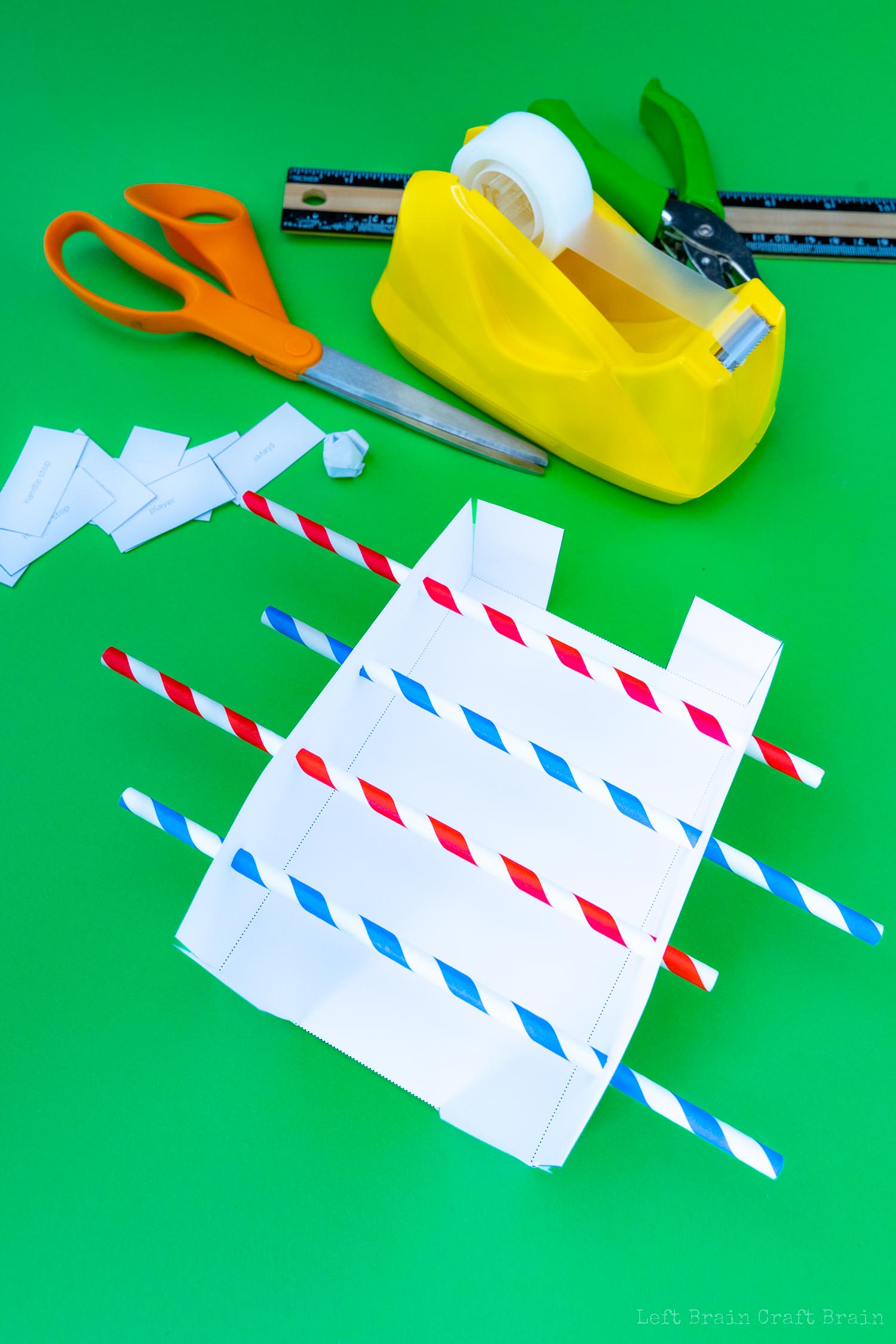 Insert straws through holes in sides of paper foosball template (green hole punch, red and blue striped paper straws, orange-handled scissors, yellow tape dispenser, black and wood ruler, white paper template, crumpled paper ball, small pieces of paper that say handle stop and player on green paper background)