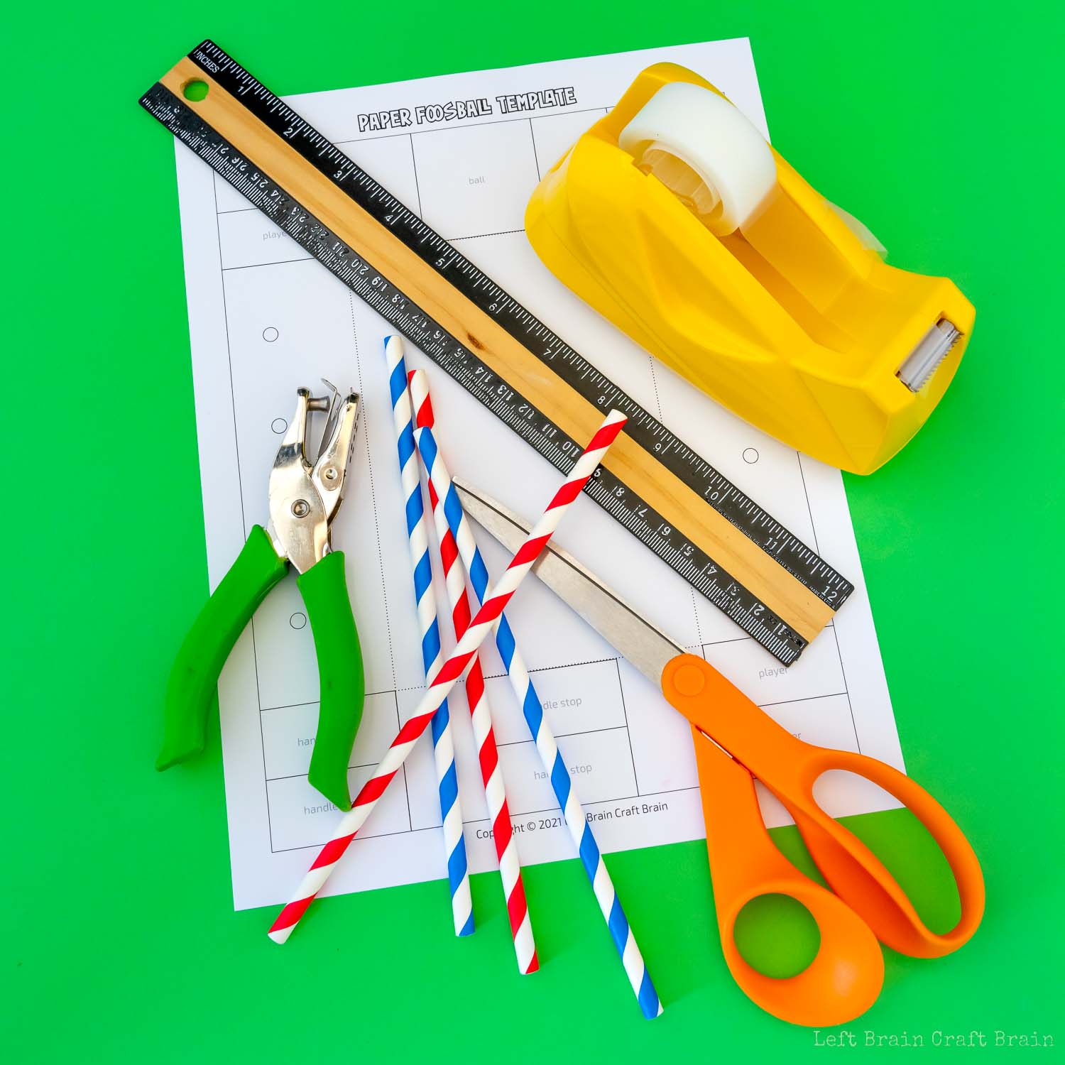 paper foosball supplies (green hole punch, red and blue striped paper straws, orange-handled scissors, yellow tape dispenser, black and wood ruler, white paper template on green paper background)
